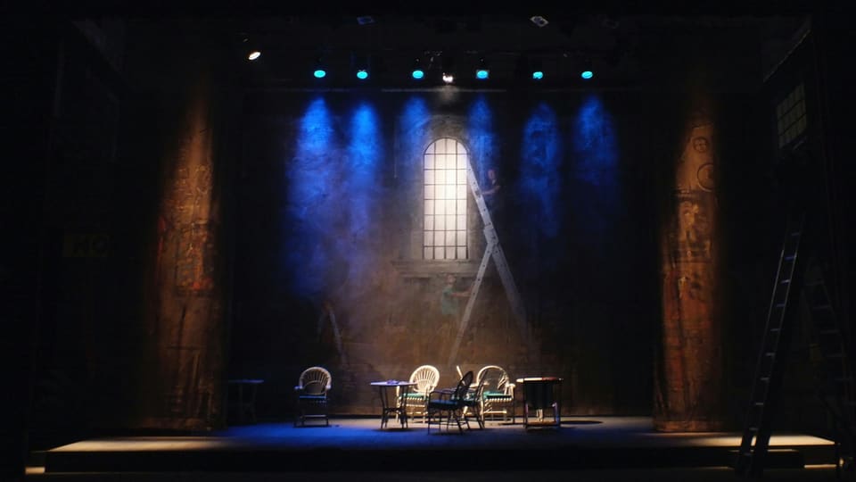 A theatre stage lit in blue, with chairs and a table